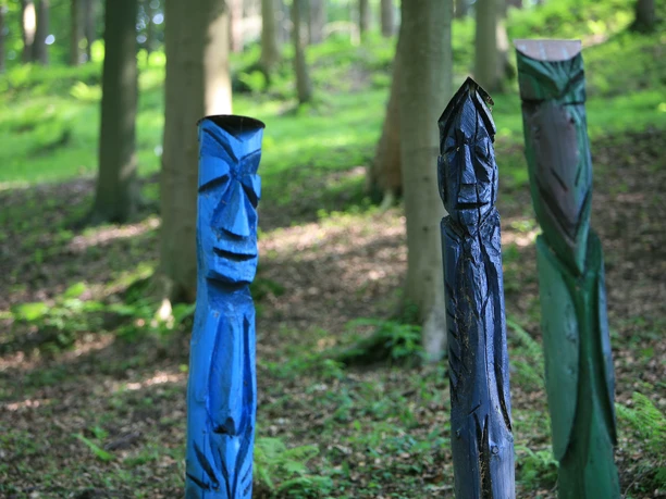 Pütz-Roth - Gärten der Bestattung Drei farbenfrohe, handgeschnitzte Totemfiguren stehen in einem sonnendurchfluteten Waldabschnitt.