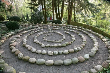Pütz-Roth - Gärten der Bestattung Ein Steinkreis in spiralförmiger Anordnung in einem bewaldeten Gartenbereich, umgeben von Bäumen.