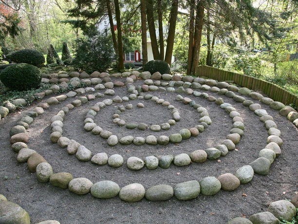 Pütz-Roth - Gärten der Bestattung Ein Steinkreis in spiralförmiger Anordnung in einem bewaldeten Gartenbereich, umgeben von Bäumen.