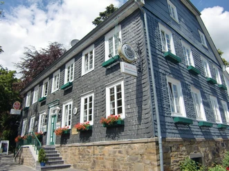 typisches Schieferhaus Historisches, graues Fachwerkhaus mit weißen Fenstern, grünen Fensterläden und blühenden Blumen.