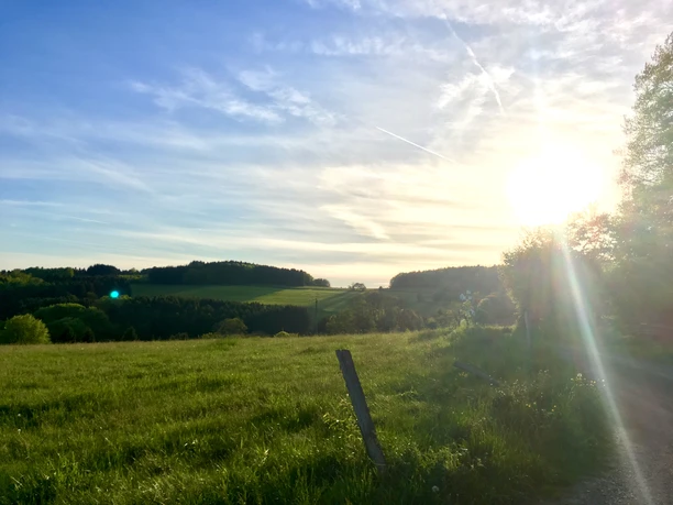 Abendstimmung Ein sonniger Tag in einer grünen Hügellandschaft mit Wäldern und einem lichten Himmel im Hintergrund.