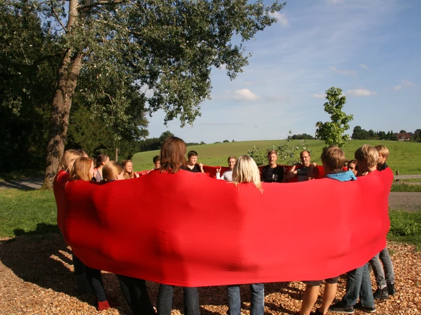 Hochseilgarten K1 Eine Gruppe von Menschen steht im Kreis, umhüllt von einem großen, roten Tuch, vor einer grünen Wiese.