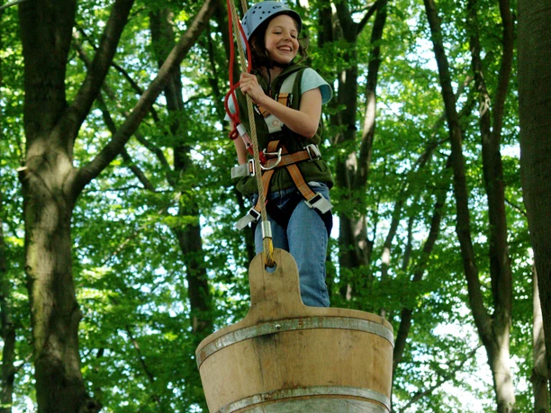 Hochseilgarten K1 Lächelndes Kind im Klettergurt schwebt über einem Fass im Abenteuerpark, umgeben von grünen Bäumen.