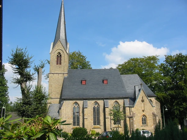 Kirche St. Agatha Steinerne Kirche mit spitzem Turm, umgeben von Bäumen, unter klarem, blauem Himmel.