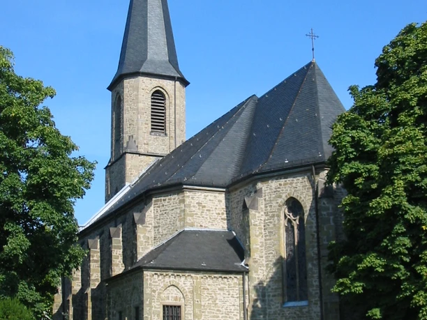 Kirche St. Johannes Apostel und Evangelist Eine steinerne Kirche mit Spitzdach und Turm, umgeben von grünen Bäumen unter klarem Himmel.