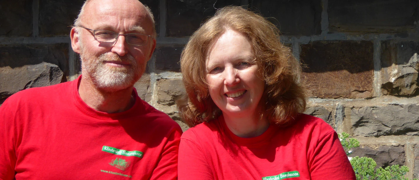 Klosterhof Bünghausen Zwei lächelnde Erwachsene in roten T-Shirts sitzen an einem Tisch vor einer Steinmauer, Sonnenschein.