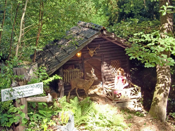 Märchenwald Altenberg Ein rustikales Holzhaus im Wald mit einem Reh und einer Schaufensterpuppe in Tracht vor der Veranda.