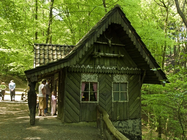 Märchenwald Altenberg Kleines, verwunschenes Holzhaus im Wald, mit Menschen davor im Sonnenschein.