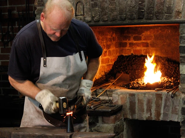 Schmied Ein Schmied in Arbeitskleidung bearbeitet glühendes Metall vor einem hell brennenden Schmiedefeuer.