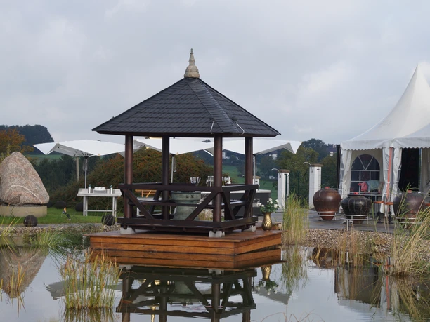 Asiatisches Museum Ein Holzpavillon auf einem schwimmenden Podest in einem Teich, umgeben von Zelten und Dekorationen.
