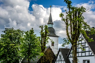 Marienberghausen Kirchturm mit Wetterhahn ragt zwischen weißen Fachwerkhäusern und grünen Laubbäumen hervor.