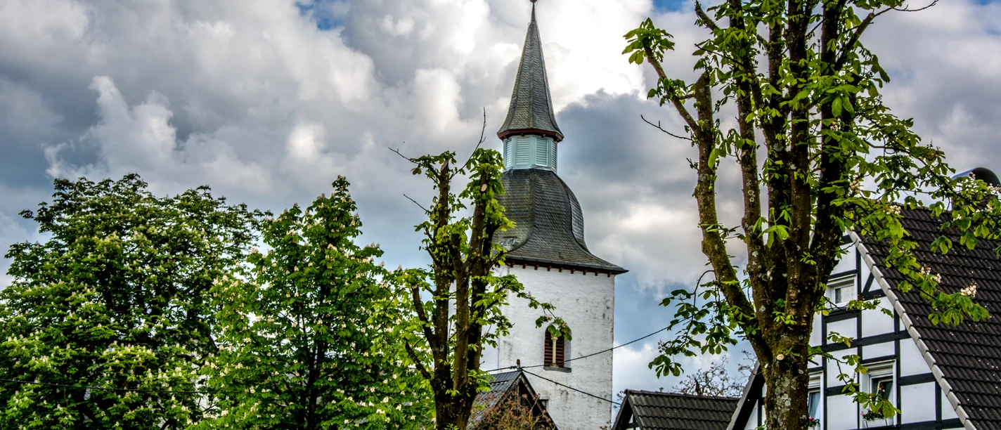 Marienberghausen Kirchturm mit Wetterhahn ragt zwischen weißen Fachwerkhäusern und grünen Laubbäumen hervor.