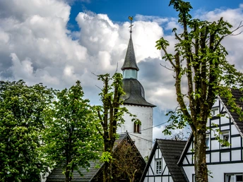Marienberghausen Kirchturm mit Wetterhahn ragt zwischen weißen Fachwerkhäusern und grünen Laubbäumen hervor.