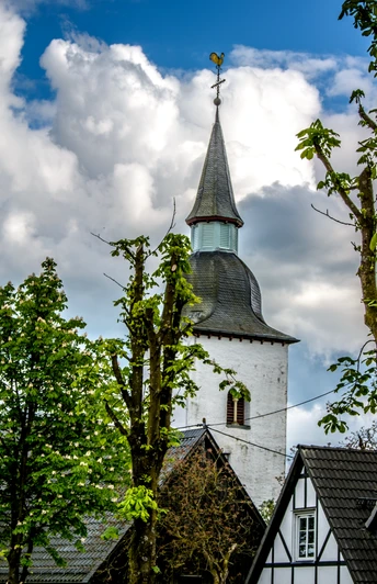 Marienberghausen Kirchturm mit Wetterhahn ragt zwischen weißen Fachwerkhäusern und grünen Laubbäumen hervor.