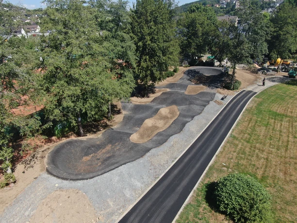 Pumptrack in Morsbach Luftaufnahme eines neu angelegten Pumptracks in einer grünen Parklandschaft, umgeben von Bäumen.