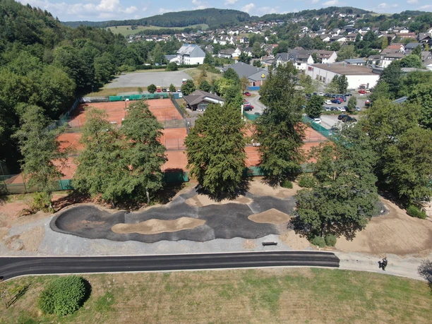 Pumptrack in Morsbach Luftaufnahme eines Sportkomplexes mit Tennisplätzen, umgeben von Bäumen in einer ländlichen Gegend.