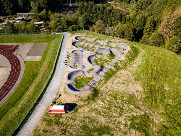 Pumptrack in Reichshof Eckenhagen Pumptrack mit geschwungenen Asphaltbahnen inmitten grüner Landschaft, umgeben von Wald und Sportplatz.