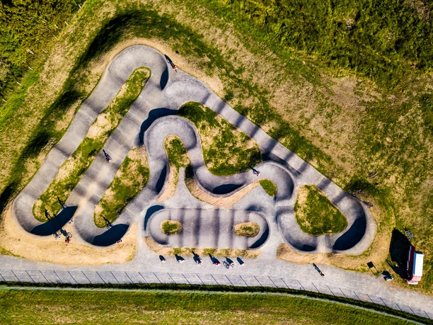 Pumptrack in Reichshof Eckenhagen Luftaufnahme eines gewundenen Pumptracks mit Radfahrern, umgeben von grüner Landschaft und Bäumen.