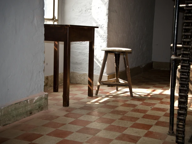 Reuschenberger Mühle Innenansicht Ein dunkler Raum mit einem hölzernen Tisch und Hocker auf einem rot-weißen Fliesenboden.