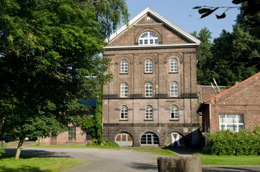 Reuschenberger Mühle Historische Ziegelsteinfassade eines vierstöckigen Gebäudes mit großen Bogenfenstern und grünem Umfeld.