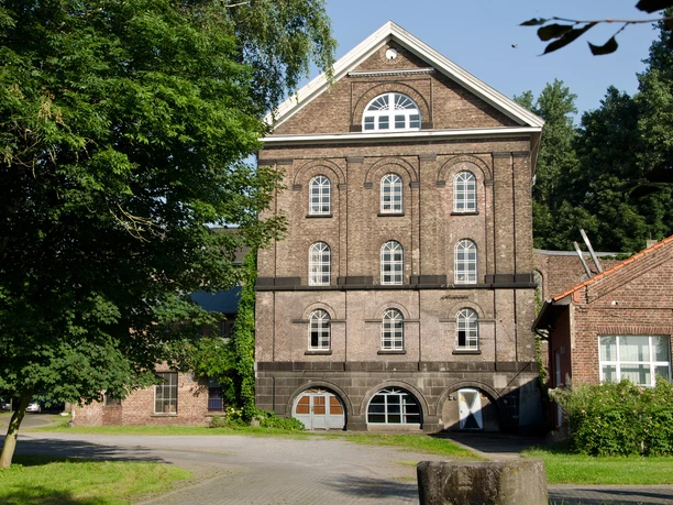 Reuschenberger Mühle Historische Ziegelsteinfassade eines vierstöckigen Gebäudes mit großen Bogenfenstern und grünem Umfeld.