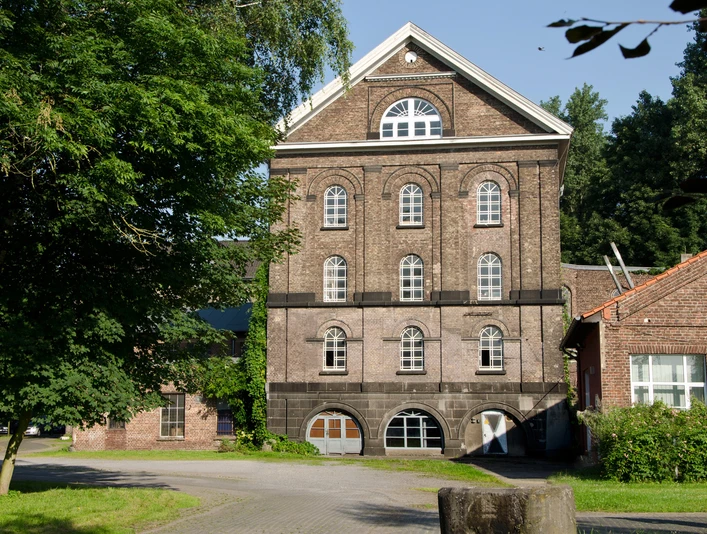 Reuschenberger Mühle Historische Ziegelsteinfassade eines vierstöckigen Gebäudes mit großen Bogenfenstern und grünem Umfeld.