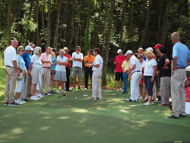 Short Game Area Eine Gruppe von Golfern steht in sportlicher Kleidung auf einem Übungsgrün vor einem Wald.