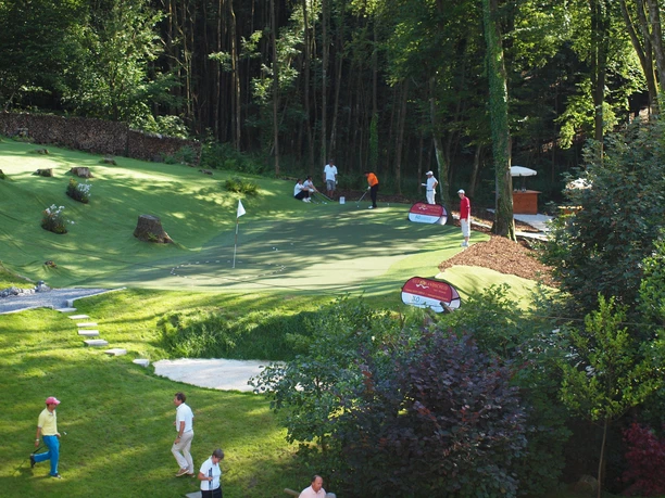 Short Game Area Mehrere Personen spielen auf einer gepflegten, grünen Minigolfanlage, umgeben von Bäumen.