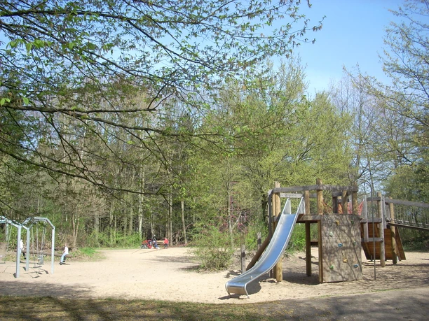 Mediendatei Spielplatz mit Klettergerüst, Rutsche und Schaukeln in parkähnlicher Umgebung, umgeben von Bäumen.