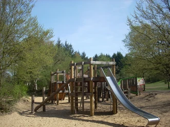 Mediendatei Holzspielplatz mit Klettergerüst, Rutsche und Sandboden, umgeben von Bäumen unter blauem Himmel.