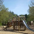 Mediendatei Holzspielplatz mit Klettergerüst, Rutsche und Sandboden, umgeben von Bäumen unter blauem Himmel.