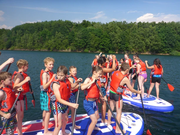 Stand-up-Paddling Kinder in Schwimmwesten paddeln auf Stand-Up-Boards in einem See, umgeben von Wald.