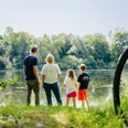 Eine Familie steht am bewachsenen Uferrand und blickt auf einen von Bäumen umgebenen See.