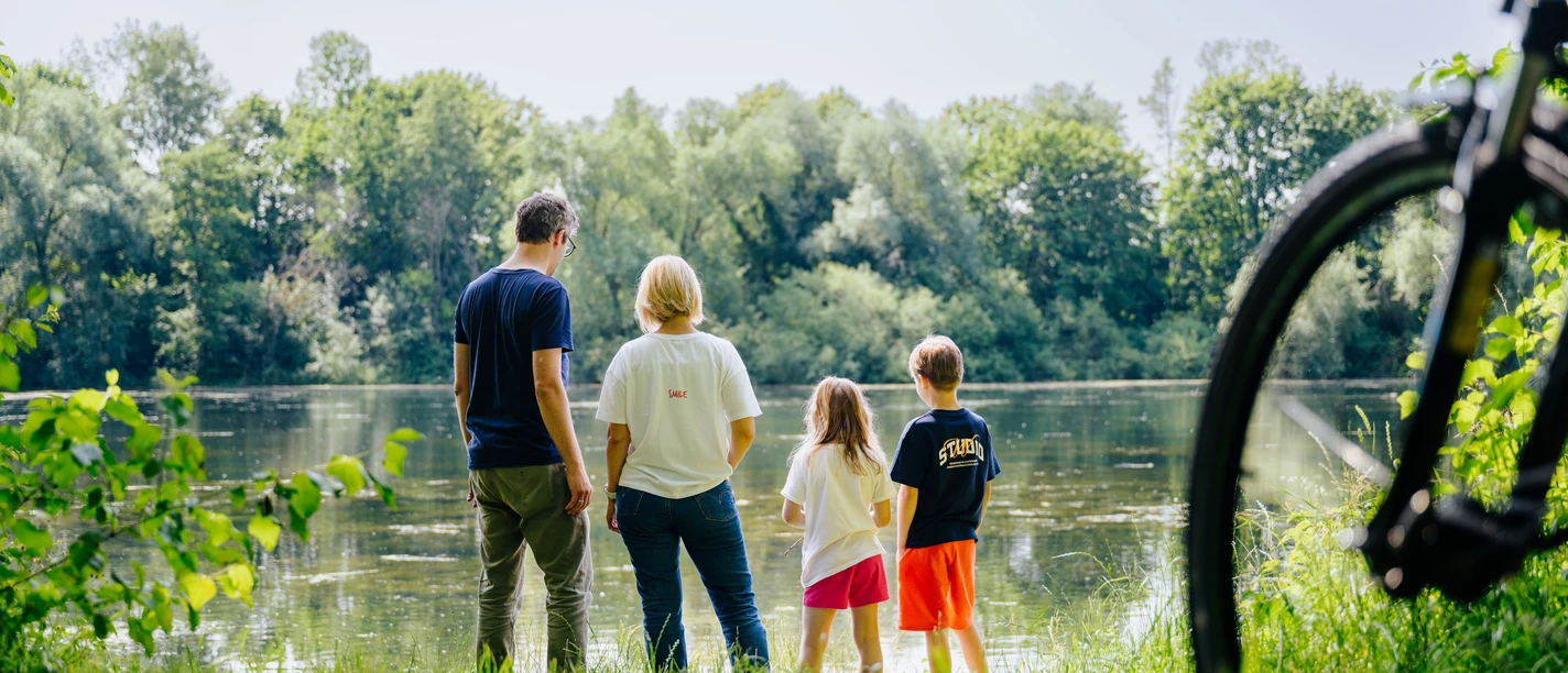 Eine Familie steht am bewachsenen Uferrand und blickt auf einen von Bäumen umgebenen See.