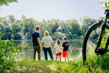 Haverkampsee_besimmazhiqi-99.jpg Eine Familie steht am bewachsenen Uferrand und blickt auf einen von Bäumen umgebenen See.