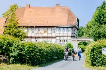 Personen, darunter Kinder auf Fahrrädern, fahren eine Straße entlang vor einem ehemaligen Jagdschloss.