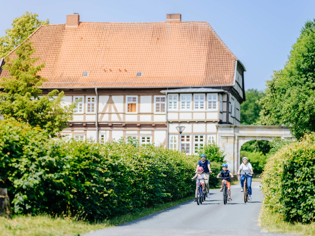 Personen, darunter Kinder auf Fahrrädern, fahren eine Straße entlang vor einem ehemaligen Jagdschloss.