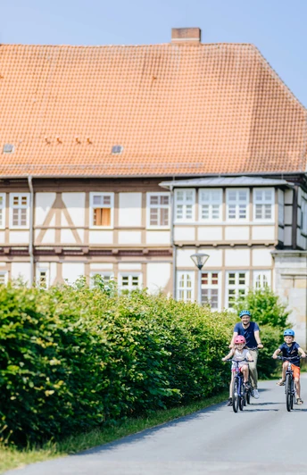 Personen, darunter Kinder auf Fahrrädern, fahren eine Straße entlang vor einem ehemaligen Jagdschloss.