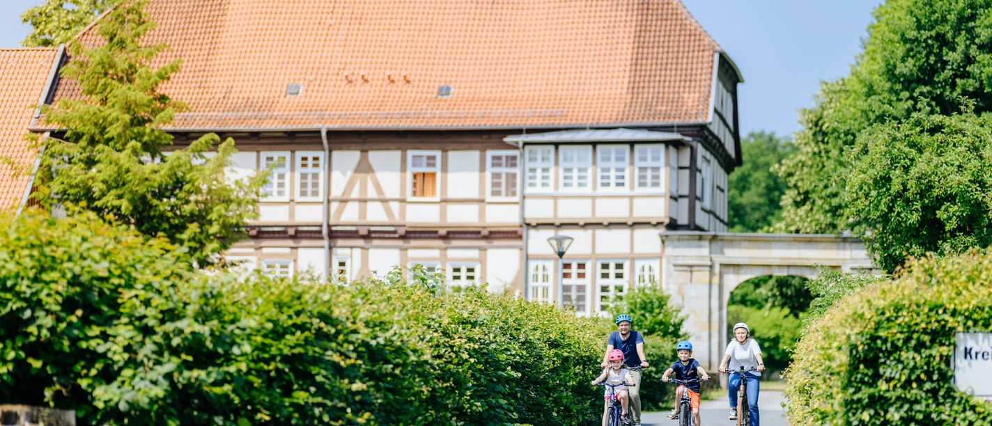 ehemaliges Jagdschloss Personen, darunter Kinder auf Fahrrädern, fahren eine Straße entlang vor einem ehemaligen Jagdschloss.