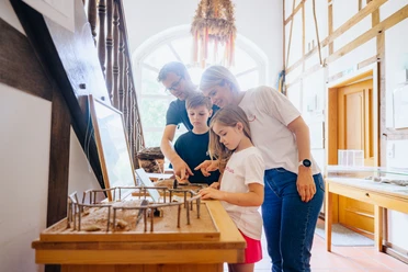 230626_besimmazhiqi-61.jpg Eine Familie betrachtet ein Miniaturmodell eines prähistorischen Hauses in einem hellen Museumsraum.
