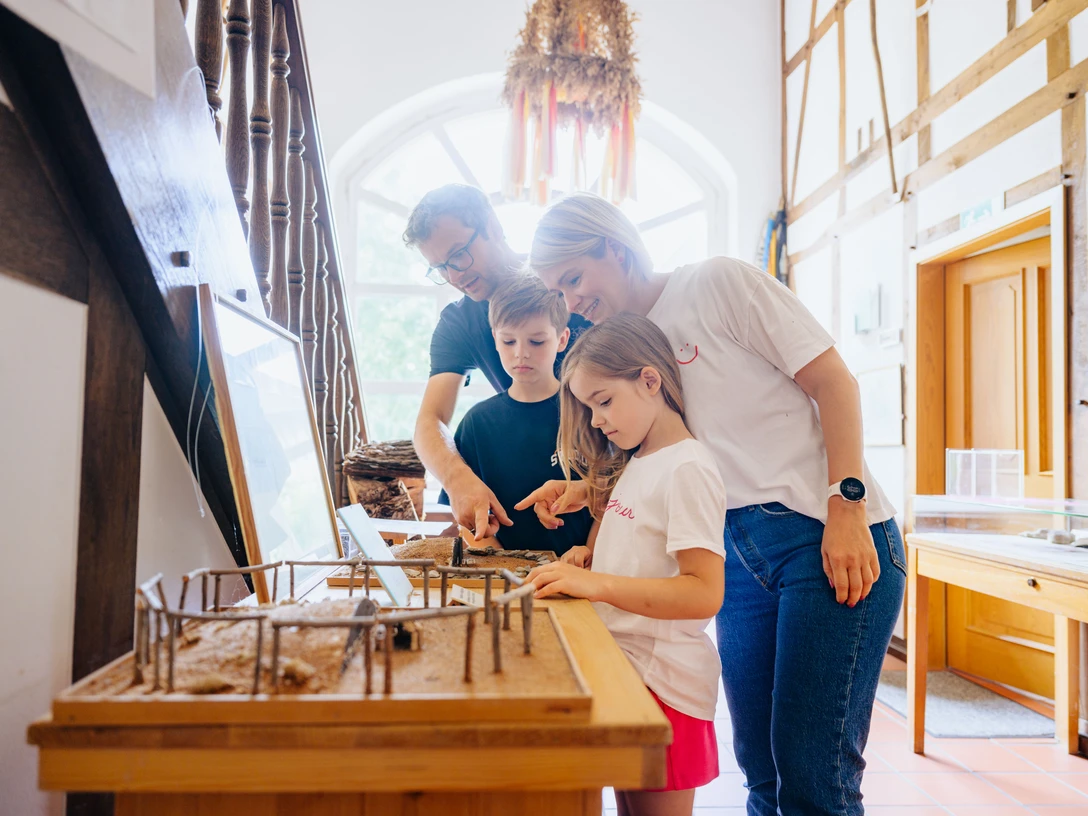 230626_besimmazhiqi-61.jpg Eine Familie betrachtet ein Miniaturmodell eines prähistorischen Hauses in einem hellen Museumsraum.