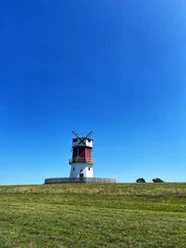 Foto Winning Mühle.jpeg Eine rote Windmühle steht auf einem grünen Hügel unter einem strahlend blauen Himmel.