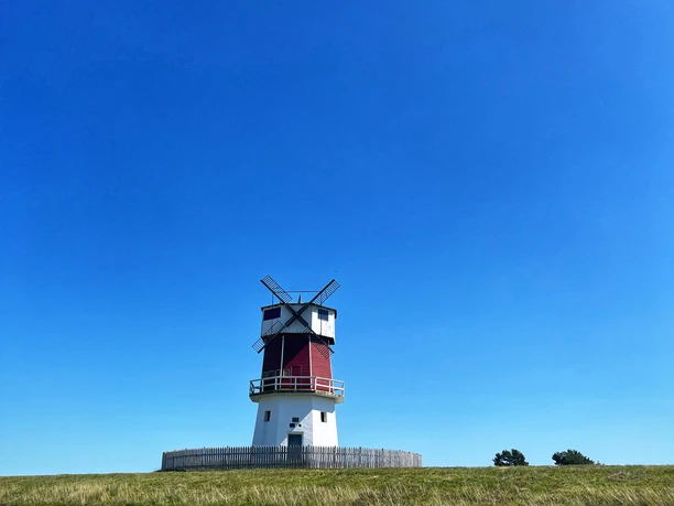 Foto Winning Mühle.jpeg Eine rote Windmühle steht auf einem grünen Hügel unter einem strahlend blauen Himmel.