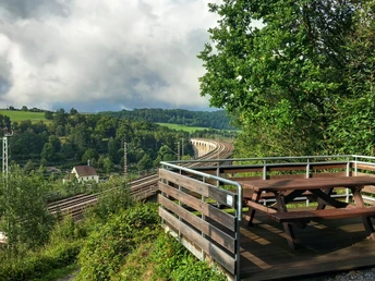 Eine Holzplattform mit Picknicktisch bietet Blick auf einen Viadukt und umliegende grüne Hügel.