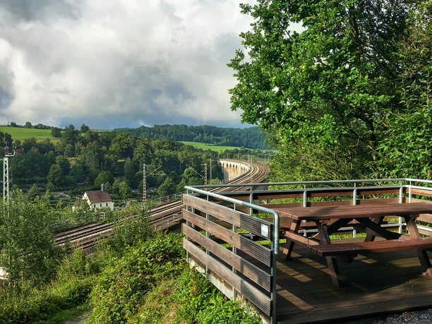 Foto Aussichtsplattform Viadukt.jpeg Eine Holzplattform mit Picknicktisch bietet Blick auf einen Viadukt und umliegende grüne Hügel.