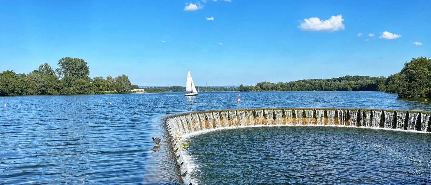 Foto Lippesee.jpeg Segelboot gleitet auf dem sonnenbeschienenen Lippesee nahe eines runden, künstlichen Wasserfalls.