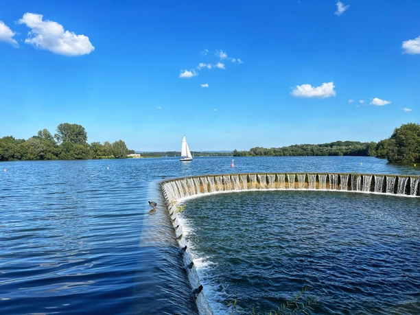 Foto Lippesee.jpeg Segelboot gleitet auf dem sonnenbeschienenen Lippesee nahe eines runden, künstlichen Wasserfalls.