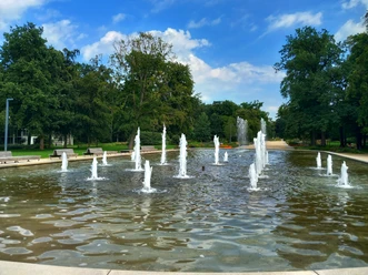 Foto Gartenschau BaLi.jpeg Wasserfontänen sprudeln lebhaft aus einem flachen Becken in einem weitläufigen, grünen Park.