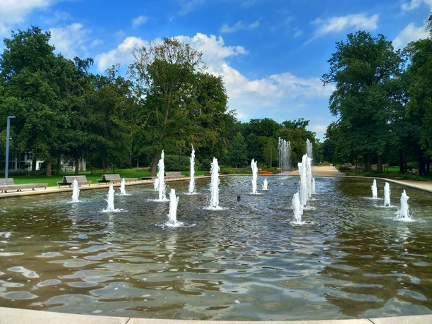 Wasserfontänen sprudeln lebhaft aus einem flachen Becken in einem weitläufigen, grünen Park.