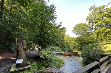 Foto Christophorus Statue.jpeg Eine hölzerne Christophorus-Statue steht am Waldesrand nahe einem sprudelnden Bach in grüner Umgebung.